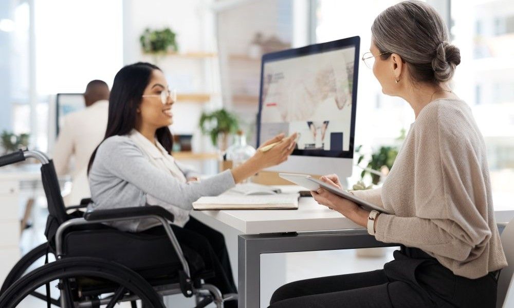 An employee who uses a wheelchair at a desk talking with a coworker.
